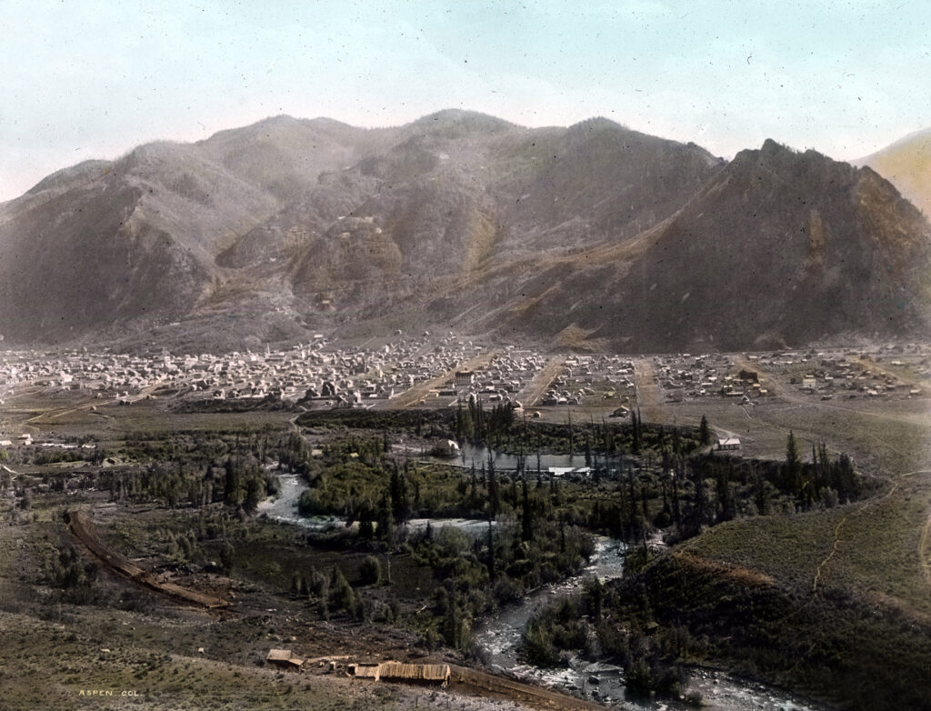 One lantern slide, colorized, of a panoramic view of Aspen taken from Red Mountain. It shows Aspen Mountain with a perfect outline of the Silver Queen (Shadow Mountain), the town, and the Roaring Fork River. Can see the D&RG Railroad bed being constructed in the foreground. Hallam Lake, streets, and west end are very clear. Taken in 1887, since the construction of the railroad tracks is visible. The D&RGRR arrived in Aspen in late Oct. 1887.